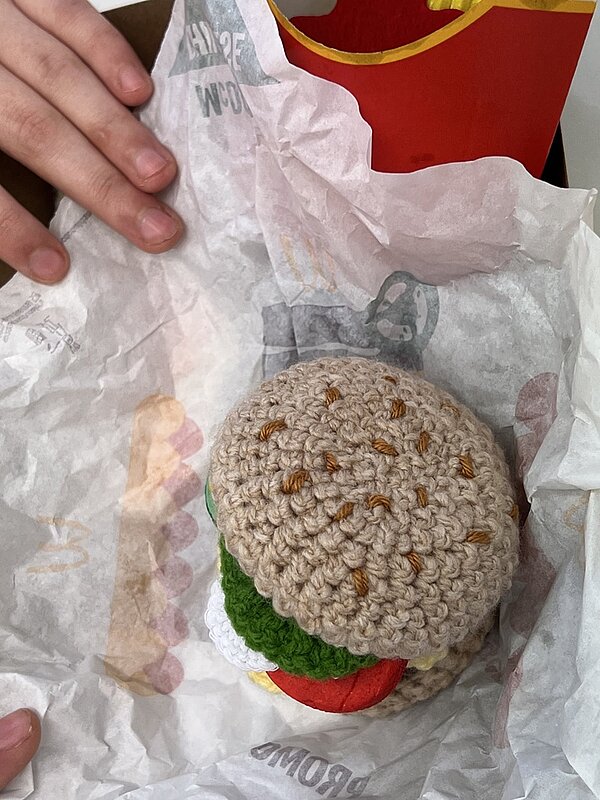  Hände packen einen in Papier eingeschlagenen, selbst gestrickten Hamburger aus.