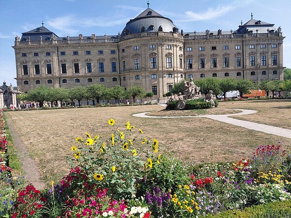 Ansicht der Würzburger Residenz mit seinen barocken Fassaden und Fenstern.  Im Vordergrund blühen bunte Blumenbeete. Der Himmel ist klar blau und die Sonne scheint.