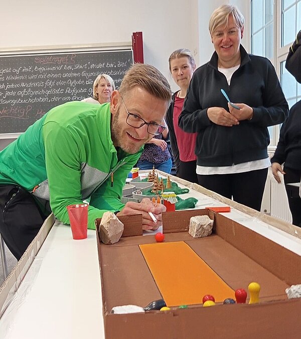 Aus Pappe, Karton und weiteren Materialen selbst gestalteter Mini-Parcours, der auf einem Tisch steht, um ihn herum die Auszubildenden und Lehrkräfte der Euro Akademie. Eine Lehrkraft hält einen Strohhalm in der Hand, um diesen als Miniqueue zu benutzen und die am Ende des Parcours aufgestellten Miniaturkegel umzustoßen.