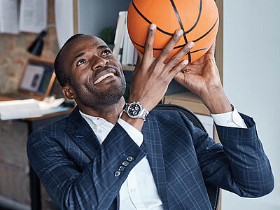 Ein Mann in einem Anzug hält einen Basketball in der Hand und bereitet sich darauf vor, ihn zu werfen. Er trägt eine Armbanduhr und steht in einem Büro mit Regalen und Papieren im Hintergrund. Der Mann lächelt und scheint sich auf den Wurf zu konzentrieren.