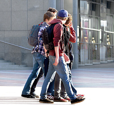 Eine Gruppe von vier Jugendlichen mit Rucksäcken läuft gemeinsam über einen sonnigen Platz vor einem modernen Gebäude. Sie tragen lässige Kleidung wie Jeans, Kapuzenpullover und Hemden und wirken im Gespräch vertieft.
