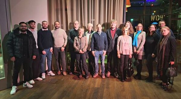 Weihnachtsfeier mit Rückblick Eine Gruppe von 16 Personen steht in einem Restaurant vor einem Hintergrund mit Vorhängen und dezenter Beleuchtung. Die Personen sind in lockerer Kleidung und lächeln in die Kamera. Einige tragen Brillen, Schals oder Taschen. Im Hintergrund ist ein beleuchtetes Schild mit dem Wort "WOW" zu erkennen. Der Boden besteht aus Holz und die Umgebung wirkt einladend.