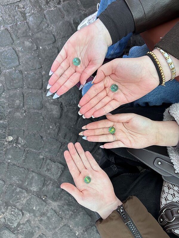 Vier Hände halten jeweils eine grüne Glasmurmel über einem gepflasterten Boden.