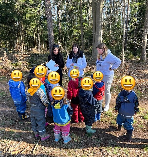 Drei angehende Sozialassistentinnen stehen im Wald vor einer Gruppe Kita-Kinder, deren Gesichter durch große Emoji-Smileys verdeckt sind. Die Kinder tragen bunte Outdoor-Kleidung und hören aufmerksam zu. Die jungen Frauen halten Arbeitsblätter in der Hand und erklären etwas.