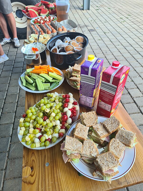 DRK Werkstätten Geithain - Aktionstag gesunde Ernährung Zu sehen ist ein Tisch, auf dem verschiedene Nahrungsmittel gestellt wurden. Ganz vorne ein Teller mit Sandwiches aus Schinken, Käse und Salat, dahinter Trauben-Käse-Spieße, daneben stehen zwei Fruchtsäfte, dahinter ein Teller mit Rohkost aus Gemüse und wiederum dahinter eine Schüssel mit Quark und eine Schüssel mit Schokomuffins.