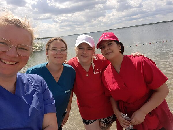 Ein Sinneskreis am Goitzsche-Strand: Lernen, Fühlen, Erleben! Vier Frauen stehen am Ufer eines Sees, lächeln in die Kamera. Alle tragen bunte T-Shirts, im Hintergrund das Wasser und der Himmel mit Wolken.