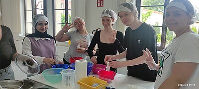 Die angehenden Kinderpflegerinnen stehen in der Küche an einem großen Tisch und bereiten gesunde und bunte Snacks aus Obst und Gemüse vor. Sie tragen Hygienehauben auf dem Kopf, lächeln und sind alle gut gelaunt. 