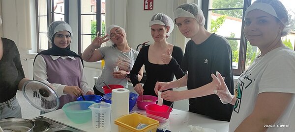 Gesund und kreativ Die angehenden Kinderpflegerinnen stehen in der Küche an einem großen Tisch und bereiten gesunde und bunte Snacks aus Obst und Gemüse vor. Sie tragen Hygienehauben auf dem Kopf, lächeln und sind alle gut gelaunt.