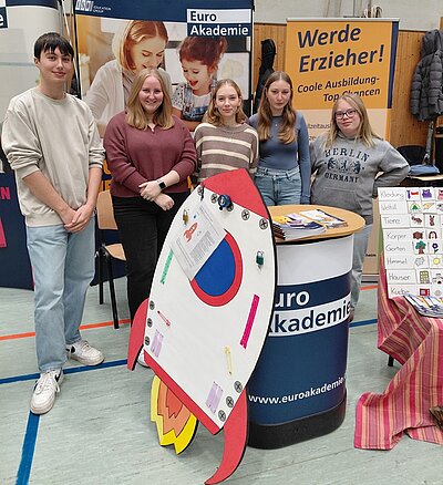 Auf dem Bild stehen fünf Auszubildende der Euro Akademie Tauberbischofsheim eng beieinander vor einem Messestand auf einer Ausbildungsmesse. Hinter ihnen befinden sich Roll-Ups mit dem Logo der Euro Akademie sowie dem Slogan ‚Werde Erzieher!‘. Vor der Gruppe steht ein großer, farbenfroher Pappaufsteller in Form einer Rakete, der für pädagogische Projekte genutzt wird. Rechts daneben liegt weiteres Anschauungsmaterial, darunter eine Tafel mit kindgerechten Symbolen zu Themen wie Kleidung, Tiere und Alltag. Auf dem runden Messtresen befinden sich Flyer und Informationsunterlagen zur Erzieherausbildung. Die Auszubildenden wirken freundlich und engagiert und repräsentieren ihre Schule in einer lebendigen Messeumgebung.