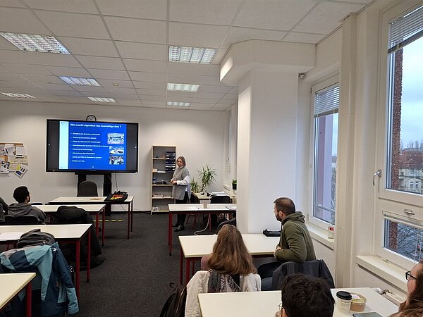 In einem Klassenzimmer zeigt eine Vertreterin des Auswärtigen Amts an einem großen Bildschirm eine Präsentation. Die Schüler hören aufmerksam zu. Das Foto wurde aus der letzten Reihe aufgenommen, so dass man die Rückenansicht der Schüler*innen sieht.