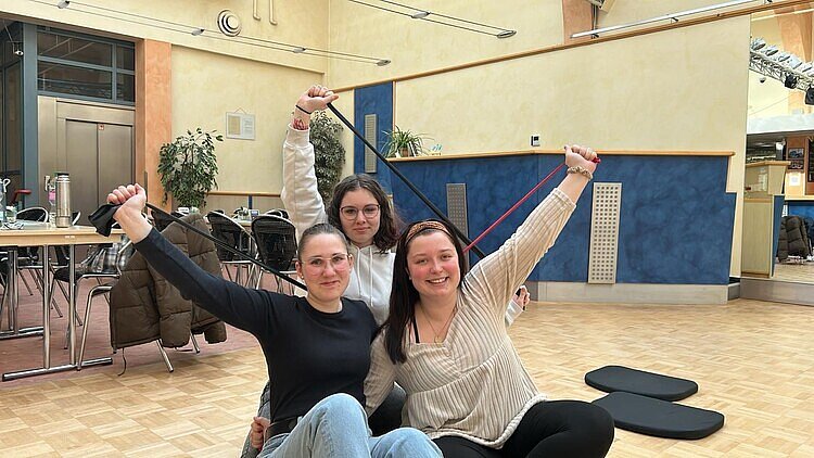 Fit für die Prüfung mit Bewegung Drei Schülerinnen haben sich für ein gemeinsames Foto in Tanzsaal positioniert: Sie halten alle drei jeweils ein Gummihand mit gestrecktem Arm fest. Im Hintergrund steht ein Tisch mit Thermoskanne darauf und über den Stühlen hängen Jacken.