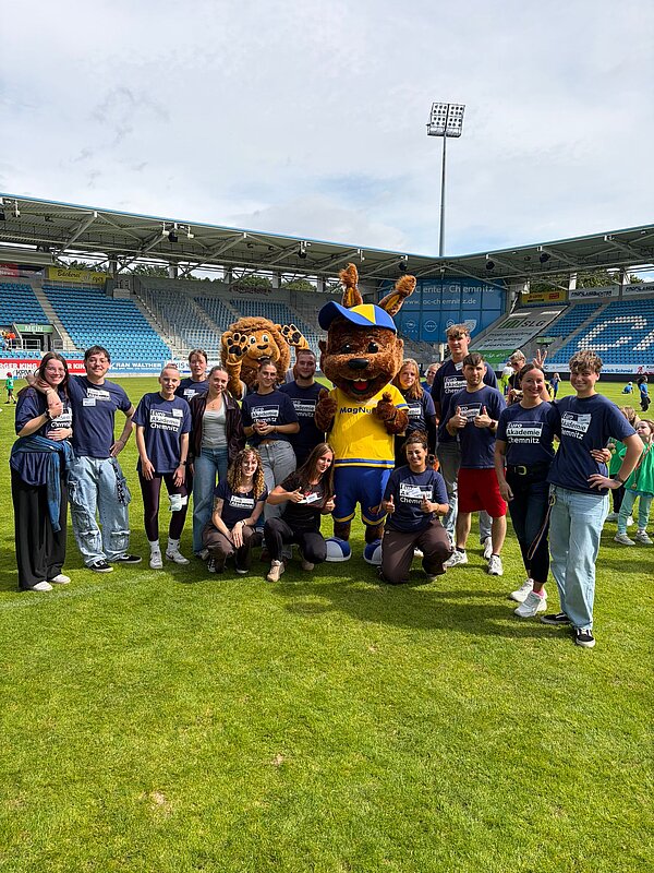 Gruppenfoto von Mitarbeitenden und Teilnehmenden der Euro Akademie Chemnitz auf dem Stadionrasen gemeinsam mit zwei großen Maskottchen.