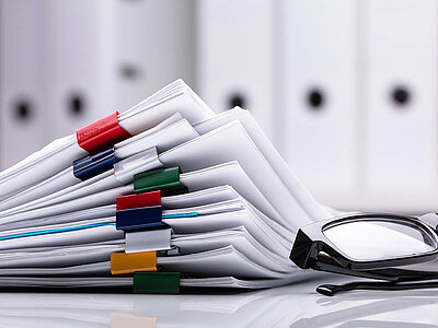 Ein Stapel Papierunterlagen, der mit bunten Büroklammern in verschiedenen Farben zusammengehalten wird. Im Vordergrund liegt eine schwarze Brille. Im Hintergrund sind verschwommene Ordner zu sehen. Das Bild symbolisiert Büroarbeit, Dokumentenverwaltung und Organisation.