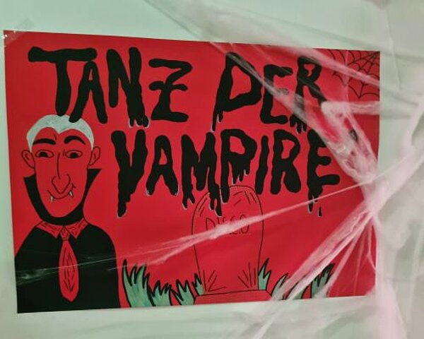 Ein großes rotes Plakat auf dem in schwarzen Buchstaben "Tanz der Vampire" geschrieben steht und ein gezeichneter Vampir zu sehen ist. über das Plakat sind Deko-Spinnweben angebracht.