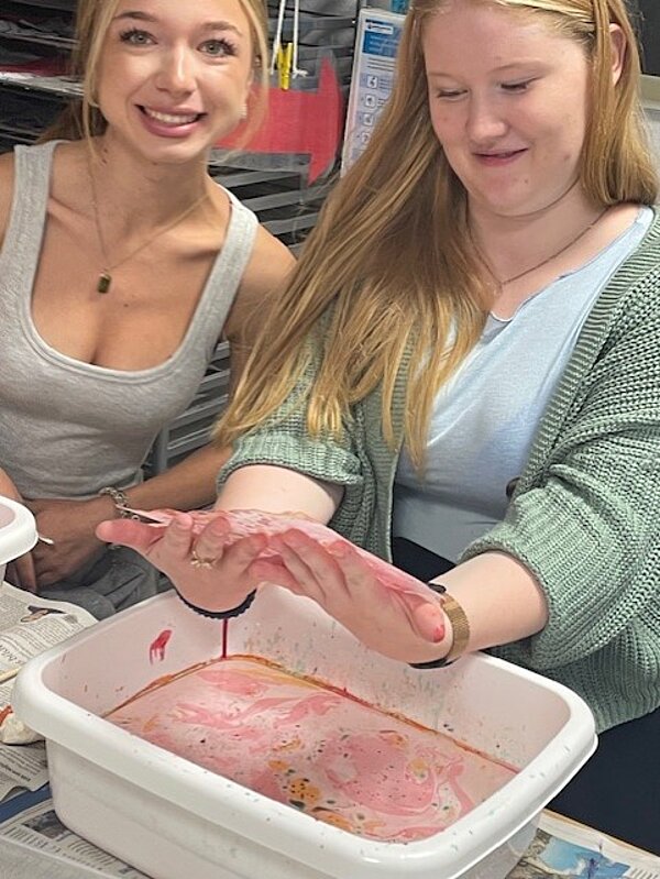 Zwei Personen sitzen an einem Tisch und arbeiten gemeinsam an einer weißen Kunststoffwanne mit roter Flüssigkeit. Die Person links, mit grauer Kleidung, lächelt in die Kamera, während die Person rechts, mit grünem Cardigan, sich auf die Wanne konzentriert. Der Tisch ist mit Zeitungen bedeckt, und im Hintergrund sind Regale zu sehen.