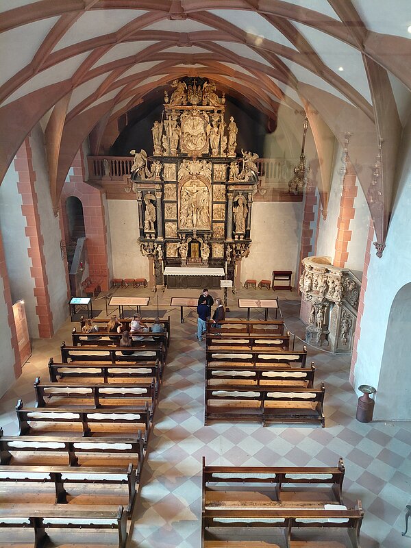 Eintauchen in die Geschichte Aschaffenburgs Blick auf das Innere einer Kirche mit hohen Decken, einem prunkvoll verzierten Altar und mehreren Reihen leerer Holzbänke. Einige Personen sitzen oder stehen im hinteren Bereich.