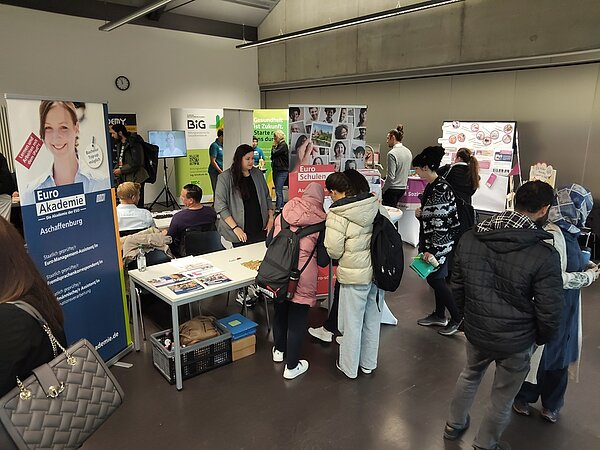 Der Blick in die Messehalle zeigt den Stand der Euro Akademie und Euro-Schulen Aschaffenburg, an der sich gerade drei junge Mädchen informieren. Am Stand steht Frau Fuchs und repräsentiert die Marken.