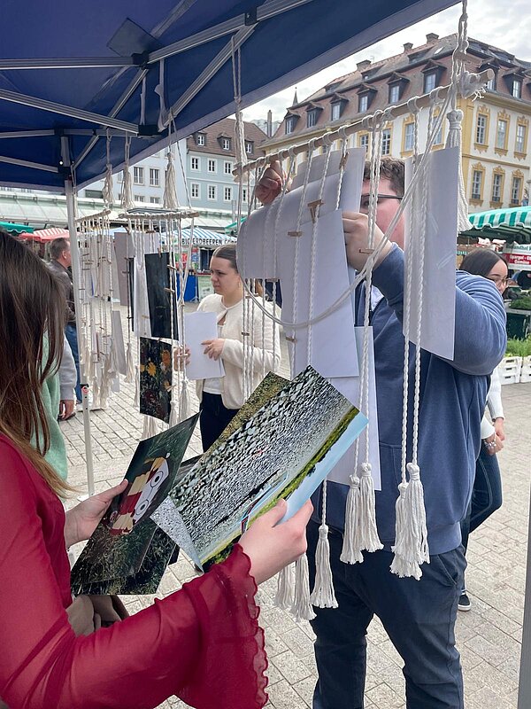 Personen hängen unter einem Pavillon Bilder und Papierblätter an eine Makramee-ähnliche Schnurwand.