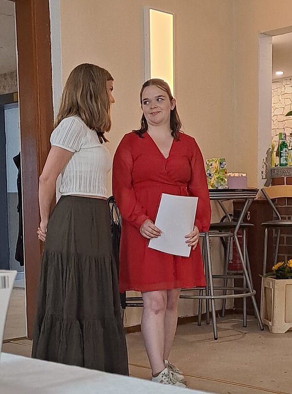 Feierliche Übergabe der Zeugnisse Zwei Mädchen grinsen sich verschmitzt an. Das rechte Mädchen trägt ein rotes Kleid und hält ein weißes Blatt in der Hand. Das Mädchen, das links steht, hat einen langen dunklen Rock an und ein helles Oberteil, sie ist nur von der Seite zu sehen.