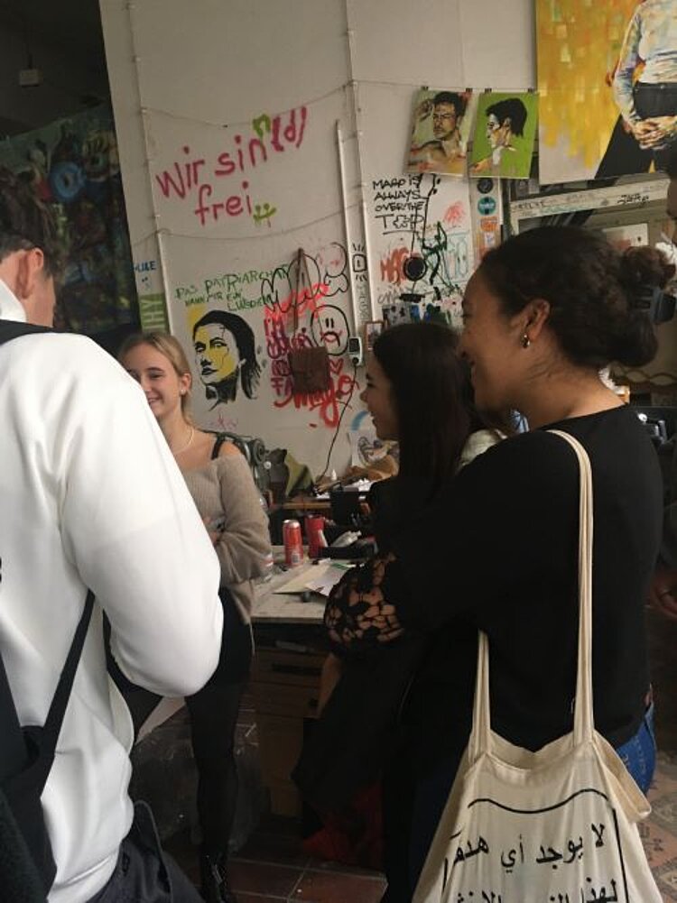 Eine Gruppe von vier Personen unterhält sich in einem künstlerisch gestalteten Atelier. Im Hintergrund sind bemalte Wände mit Graffiti und Zeichnungen zu sehen, darunter der Schriftzug „Wir sind frei“. Zwei Frauen stehen mit einer Tasche und einem Getränk in der Hand, während eine andere Person lächelt. Die Szene wirkt lebendig und kreativ.