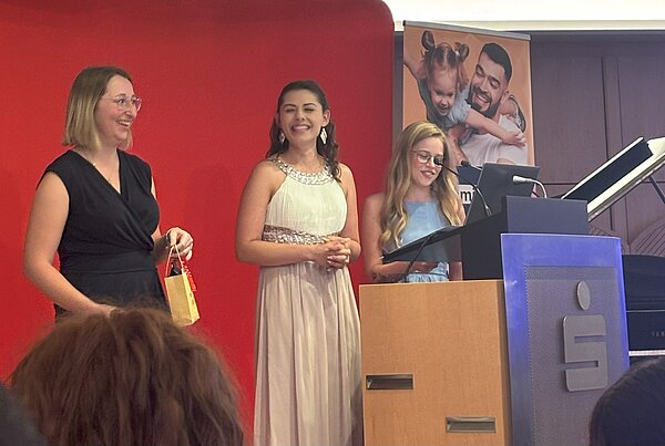 Feierlicher Abschied: Exmatrikulation der Absolvent*innen 2025 Drei Frauen stehen auf einer Bühne. Eine der Frauen steht an einem Podium und spricht. Sie lächeln und sind festlich gekleidet. Das Publikum sitzt und hört zu. Im Hintergrund ist ein rotes Banner mit einem Logo und ein Klavier zu sehen.