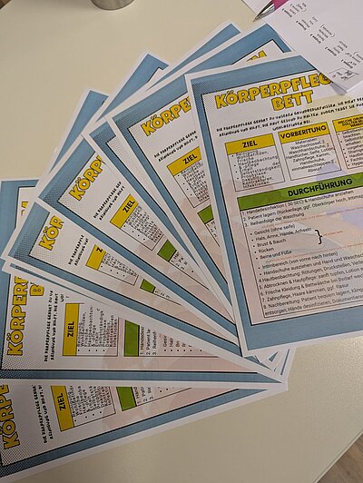 Handouts zum Thema "Körperpflege im Bett" sind auf einem Tisch aufgefächert ausgelegt.