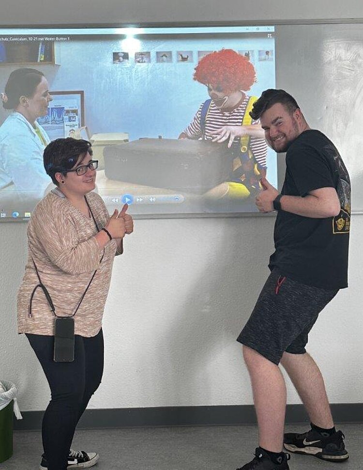 Euro Akademie erhält Auszeichnung „Clever in Sonne und Schatten“ 2024 Zu sehen ist links eine Frau mittleren Alters mit schwarzer Hose und hellbraunen Oberteil, die ihr Daumen nach oben hält. Rechts ist ein junger Mann mit kurzer schwarzer Hose und schwarzem Oberteilt, der ebenfalls seine Daumen nach oben hält. Im Hintergrund ist ein Whiteboard, auf welchem ein Film abgespielt wird oder ein Foto zu sehen ist, auf dem eine Frau mit weißem Kittel und ein Mann als Clown verkleidet einen Koffer öffnen.