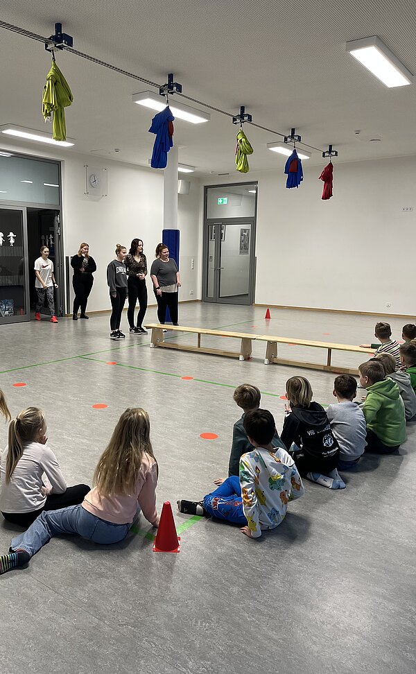 Kinder sitzen in einer Turnhalle am Boden und schauen nach vorn, wo mehrere Erwachsene an einer Bank stehen; farbige Markierungen und Hütchen liegen auf dem Boden.
