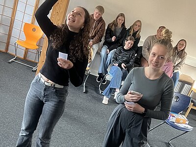 Zwei Personen stehen in einem Klassenzimmer. Eine Person steht und deutet nach oben, während sie ein Blatt Papier in der Hand hält. Die andere Person sitzt auf einem Stuhl und hält ebenfalls ein Blatt Papier. Im Hintergrund sitzen mehrere Personen auf Stühlen und beobachten die Szene.