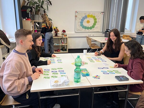 Drei Schülerinnen und ein Schüler sitzen in einem Klassenraum an einem großen Tisch, auf dem sie die Karten für das Spiel SKYJO ausgebreitet haben. Auf dem Tisch steht außerdem noch eine Plastikwasserflasche und eine Bäckertüte.