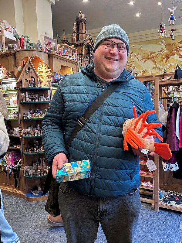 Eine Person in einem grünen Anorak und grüner Mütze steht in einem Spielzeugladen. Sie hält ein orangefarbenes Plüschtier in Form eines Fisches in der einen und eine Box mit einem Würfelspiel in der anderen Hand und lächelt dabei.