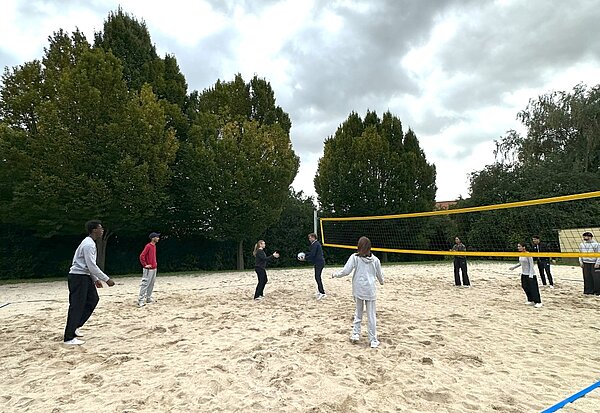 Together in motion – Bewegung, Spaß und Teamgeist Eine Gruppe von Menschen spielt auf einem Sandplatz Beach-Volleyball. Einige Spieler*innen stehen bereit, während eine Person den Ball wirft. Im Hintergrund sind Bäume und ein bewölkter Himmel zu sehen.