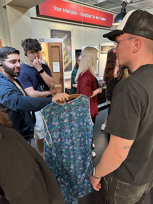 Jugendliche stehen in einer Ausstellung zusammen, ein Teilnehmer hält ein geblümtes Kleid hoch. Im Hintergrund Banner mit der Aufschrift „Unser Arbeitsplatz – Kampfplatz für den Frieden!“ sowie weitere Ausstellungselemente.