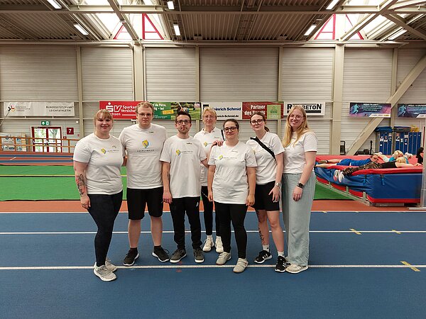 Eine Gruppe angehender Heilerziehungspfleger*innen steht gemeinsam in einer Sporthalle auf einer Laufbahn und trägt weiße Chemlympics-Shirts.