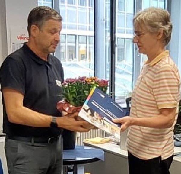 Ein Mann überreicht einer Frau einen Blumenstrauß und eine Mappe in einem Büro mit großen Fenstern.