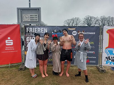 Drei Schülerinnen, ein Schüler und eine Lehrkraft, die bester Laune sind, posieren vor einem Plakat mit der Aufschrift "Frieren für den guten Zweck", natürlich in Badekleidung, teilweise notdürftig mit Bademantel und Mützen bekleidet. Die Anzeige im Hintergrund zeigt 8 Grad Lufttemperatur an.