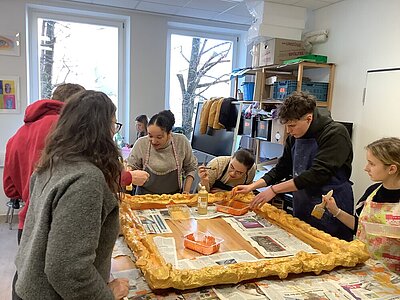 Sechs Schülerinnen stehen um einen hohen Tisch herum, auf dem ein goldener Rahmen liegt. Sie tragen Schürzen und haben Pinsel in der Hand, mit denen sie den riesigen Bilderrahmen mit Farbe bemalen.