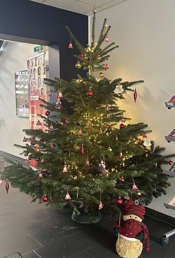 Ein geschmückter Weihnachtsbaum steht in einem Innenraum der Euro Akademie Zwickau während des Tages der offenen Tür. Der Baum ist mit kleinen roten und weißen Papierengelanhängern, roten Kugeln und warmweißen Lichterketten dekoriert. Unter dem Baum befindet sich eine große, handgefertigte Weihnachtsfigur aus Stroh, die mit einem roten Schal und einer roten Mütze bekleidet ist. Im Hintergrund sind ein schwarzer Türrahmen, ein Flur mit grauem Boden und eine Wand mit einem Plakat zu sehen. Die Atmosphäre wirkt festlich und einladend.