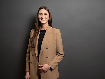 Absolventin der Euro Akademie Aschaffenburg Samira posiert vor grauem Hintergrund lächelnd in einem beigefarbenen Mantel und mit offenen langen, braunen Haaren für ein Foto.