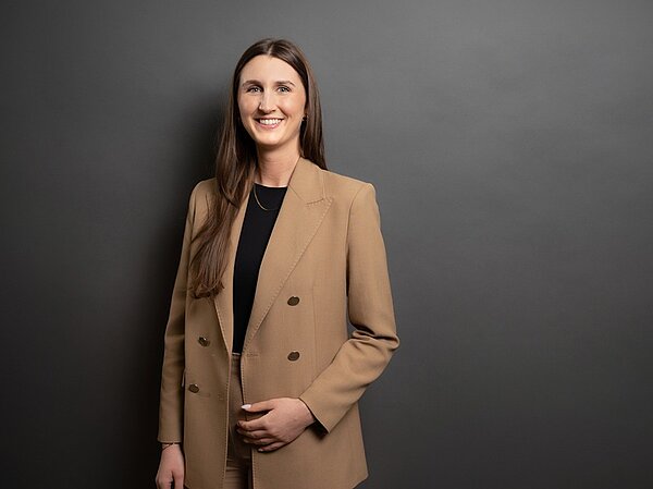 Alumni im Gespräch: Interview mit Samira Amrhein Absolventin der Euro Akademie Aschaffenburg Samira posiert vor grauem Hintergrund lächelnd in einem beigefarbenen Mantel und mit offenen langen, braunen Haaren für ein Foto.