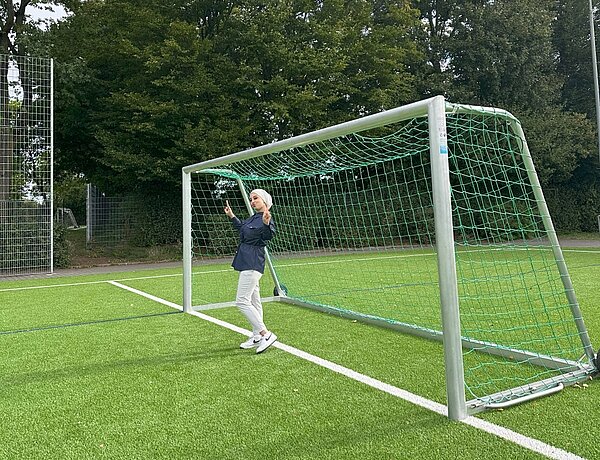 Together in motion – Bewegung, Spaß und Teamgeist Eine Person steht vor einem Fußballtor auf einem Kunstrasenplatz und macht ein Handzeichen. Die Person trägt ein Kopftuch, eine helle Hose und ein dunkelblaues Jeanshemd. Im Hintergrund sind Bäume und ein Zaun zu sehen.