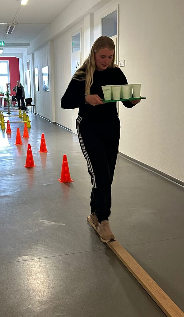 Eine junge Frau balanciert mit einem Tablett und Bechern über eine Holzlatte in einem Flur mit aufgebautem Parcours.