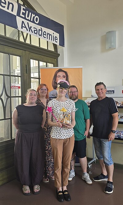 Die frisch gebackene Kauffrau für Büromanagement steht nebeneinander ihren Kolleginnen und Kollegen der Euro Akademie Halle in einem Innenraum vor einer großen Glastür und posiert für ein Erinnerungsfoto. Die nun ehemalige Azubine hält ein Geschenk mit Schleife und eine Blume in der Hand. Im Hintergrund hängt ein Banner mit einem Porträtfoto einer Frau und ein Schild mit dem Wort ' Euro Akademie'. Die Personen tragen Freizeitkleidung und blicken in Richtung Kamera.