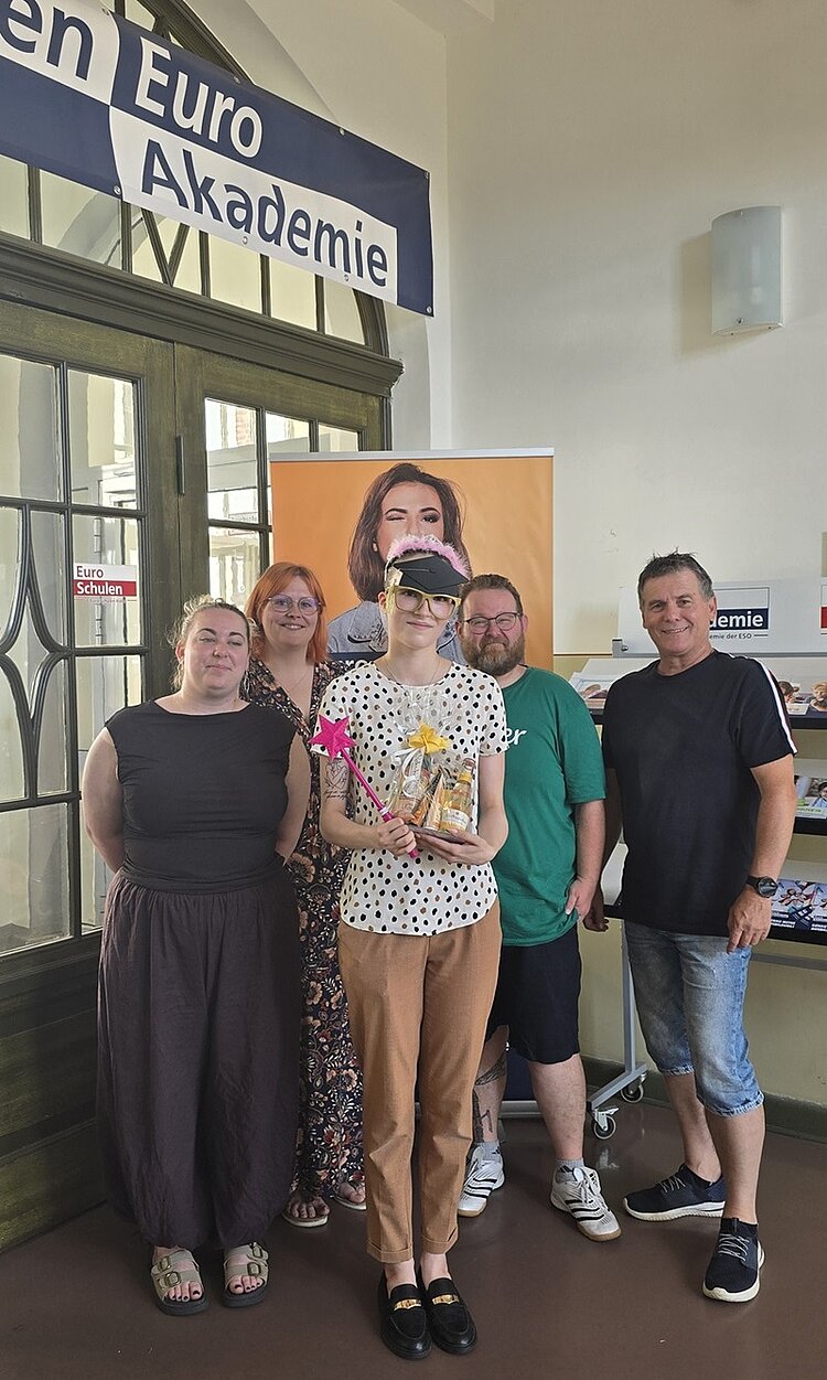 Die frisch gebackene Kauffrau für Büromanagement steht nebeneinander ihren Kolleginnen und Kollegen der Euro Akademie Halle in einem Innenraum vor einer großen Glastür und posiert für ein Erinnerungsfoto. Die nun ehemalige Azubine hält ein Geschenk mit Schleife und eine Blume in der Hand. Im Hintergrund hängt ein Banner mit einem Porträtfoto einer Frau und ein Schild mit dem Wort ' Euro Akademie'. Die Personen tragen Freizeitkleidung und blicken in Richtung Kamera.