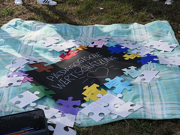 Auf einer hellblau-weiß karierten Decke liegen zahlreiche große Puzzleteile aus Papier, die mit bunten Farben bemalt und beschriftet sind. In der Mitte befindet sich eine schwarze Fläche mit weißer Schrift, auf der steht: 'Pädagogische Wertschätzung' und darunter ist ein Herz gezeichnet. Am unteren Bildrand steht ein schwarzer Kunststoffkorb mit Stiften. Die Szene spielt im Freien, das Gras und einige Schuhe sind am Rand sichtbar.