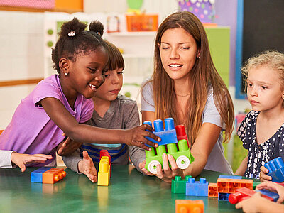 Eine Erzieherin sitzt mit drei Kindern an einem Tisch und spielt mit bunten Bauklötzen. Die Kinder wirken konzentriert und interessiert, während sie gemeinsam mit der Erzieherin ein Modell bauen. Der Raum ist hell und farbenfroh gestaltet.