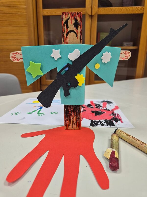 Ein Kunstwerk aus Holz und Papier: Eine Holzfigur mit einem traurigen Gesicht und einem schwarzen Gewehr, das darauf geklebt ist. Um die Figur herum sind Sterne, Herzen und Blumen aus Papier. Im Vordergrund liegt ein großer roter Handabdruck.
