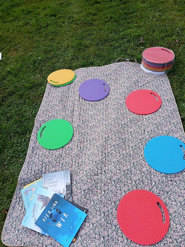 Eine graue Decke, auf der bunte runde Schaumstoffscheiben in verschiedenen Farben (rot, blau, grün, lila, gelb) liegen. Daneben liegen ein Stapel bunten Scheiben und ein Stapel mit Büchern, oben auf ein Buch mit dem Titel "Hier sind wir".