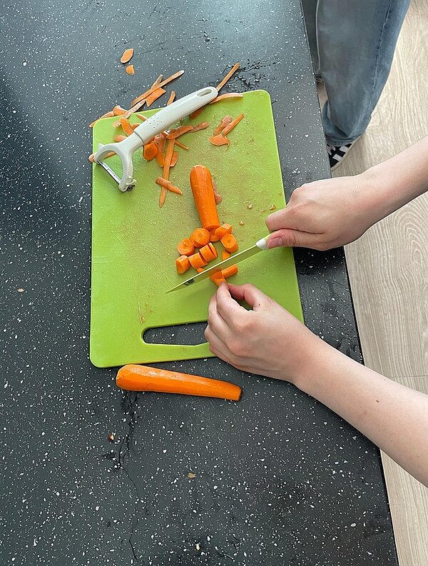 Zwei Hände schneiden eine orange Karotte auf einem grünen Schneidebrett in Scheiben. Ein Sparschäler und Karottenreste liegen ebenfalls auf dem Brett. Die Szene spielt auf einer dunklen Küchenarbeitsplatte, im Hintergrund ist ein Teil eines Beins mit heller Hose und Sportschuh zu sehen.