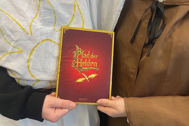 Die beiden Auszubildenden zu Kinderpfleger*innen halten gemeinsam ein Buch mit rotem Einband und goldener Verzierung. Der Titel 'Pfad der Helden' ist in goldener Schrift mittig auf dem Buchdeckel zu sehen, darunter ein goldener Pfeil. Die linke Person trägt ein weißes Gewand mit gelben Linien, die rechte Person ein braunes Gewand. Beide sind nur bis zur Brust sichtbar, Gesichter sind nicht zu erkennen.